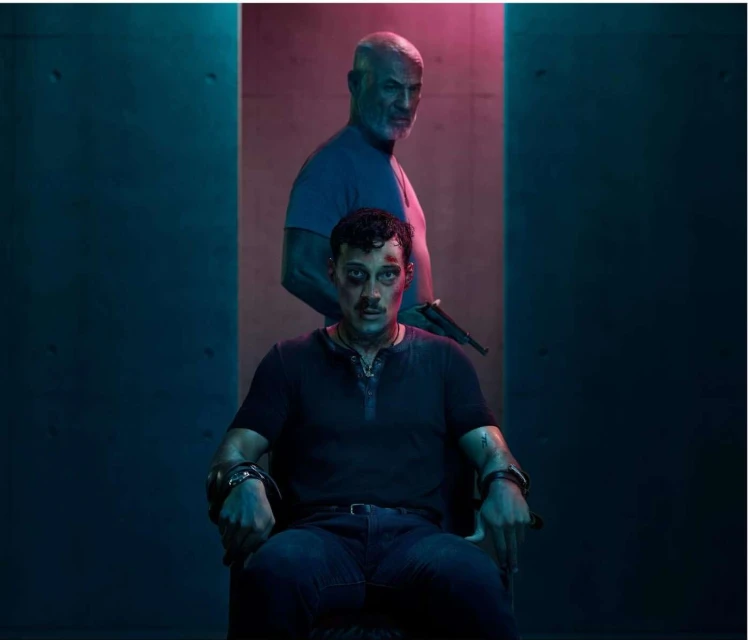 Two men in a dimly lit room. One is seated in a chair with arms bound by metal restraints; the other stands over with a gun.