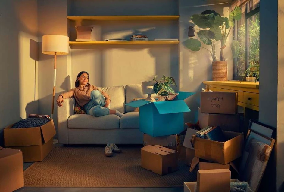 Woman lounges on a sofa surrounded by moving boxes in a warmly lit room with a large indoor plant near a sunny window.
