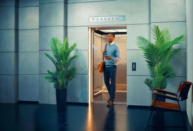 An African-American man exits a modern elevator, holding his cell phone and a leather bag, in an indoor setting with potted plants.