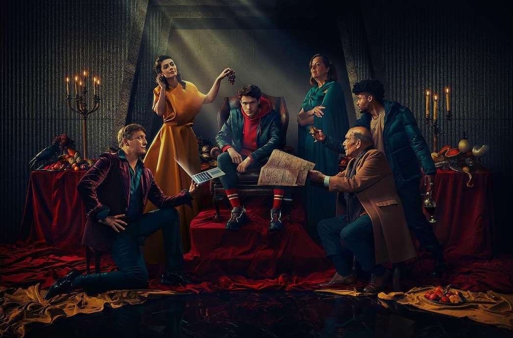 Group of people gathered indoors with dramatic lighting, featuring adults in formal wear, and tables with fruits and books.