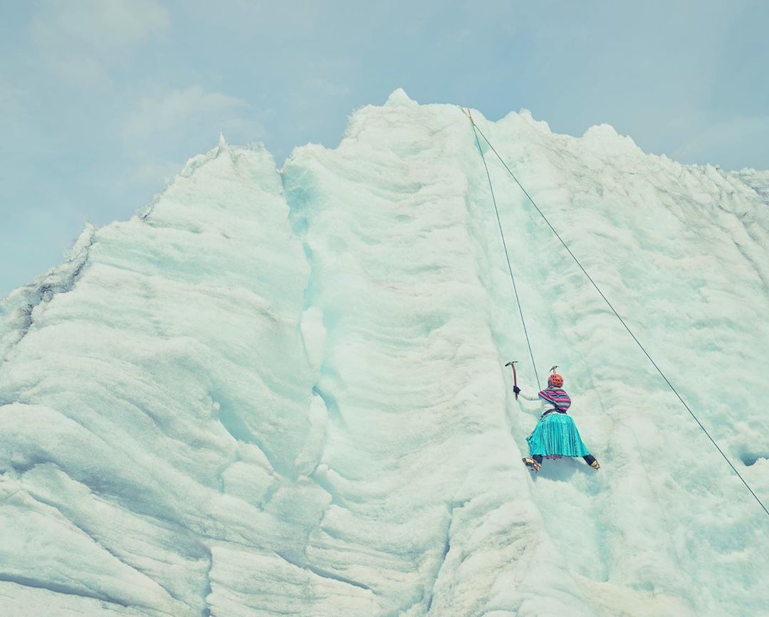 A person in a turquoise skirt climbs a glacial mountain face, using ice axes, secured by a rope against a blue sky.