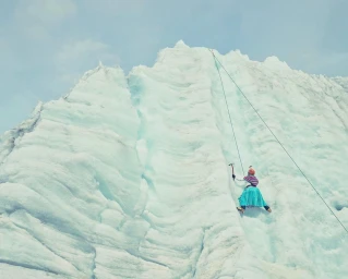 Climber in a blue skirt scaling a massive vertical glacier wall using ropes and ice axes under a bright sky.