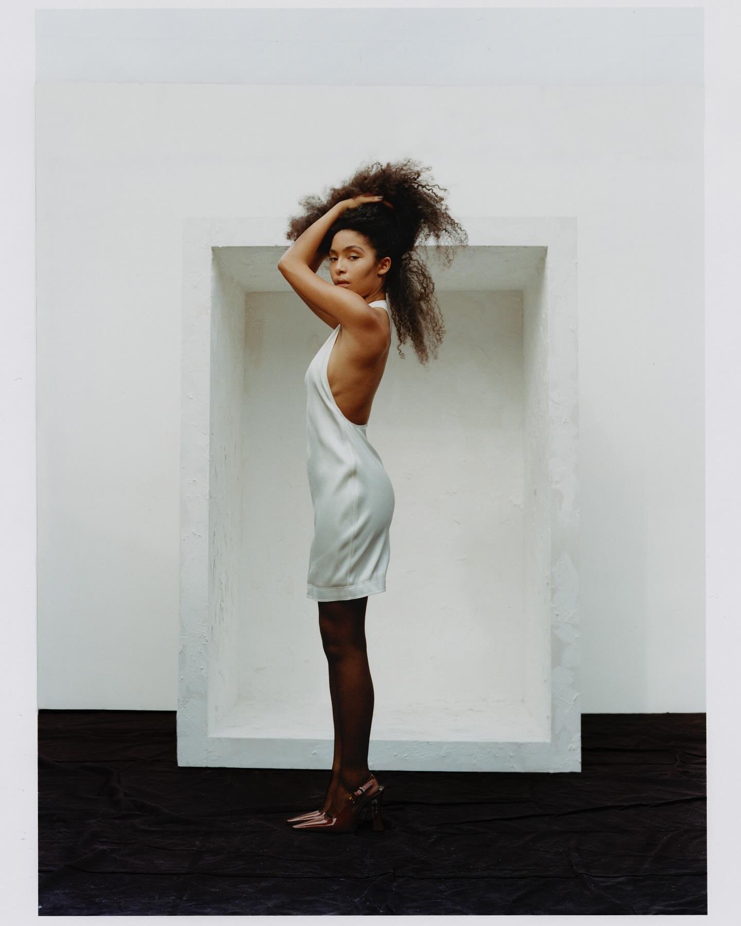 A woman with curly hair in a white dress and heels stands in profile against a white wall with a dark floor, in an indoor setting.