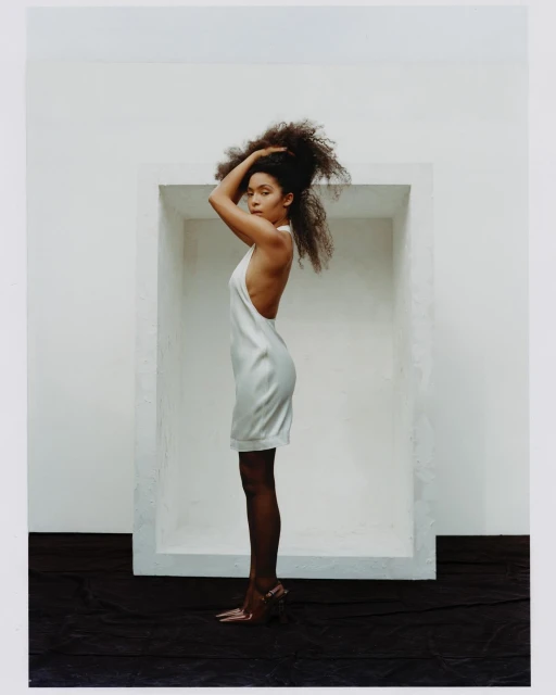 A woman with curly hair in a white dress and heels stands in profile against a white wall with a dark floor, in an indoor setting.