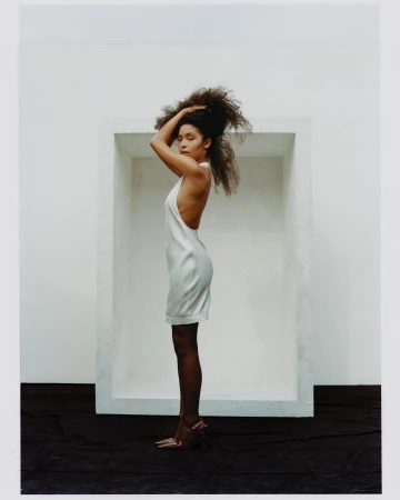 A woman with curly hair in a white dress and heels stands in profile against a white wall with a dark floor, in an indoor setting.