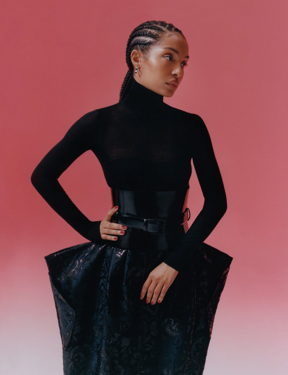 Woman in a black turtleneck and high-waisted sculptural skirt poses against a pink background in a fashion editorial shot.