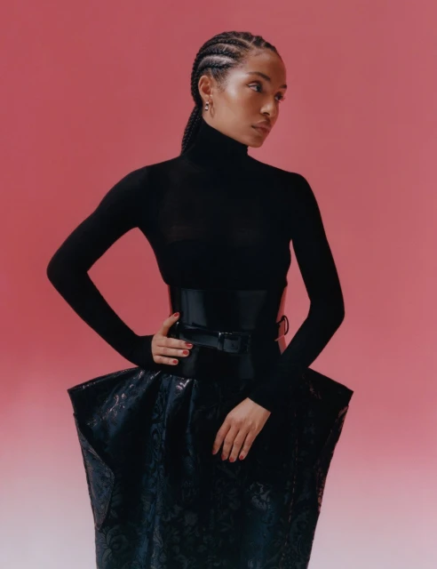 Woman in a black turtleneck and high-waisted sculptural skirt poses against a pink background in a fashion editorial shot.