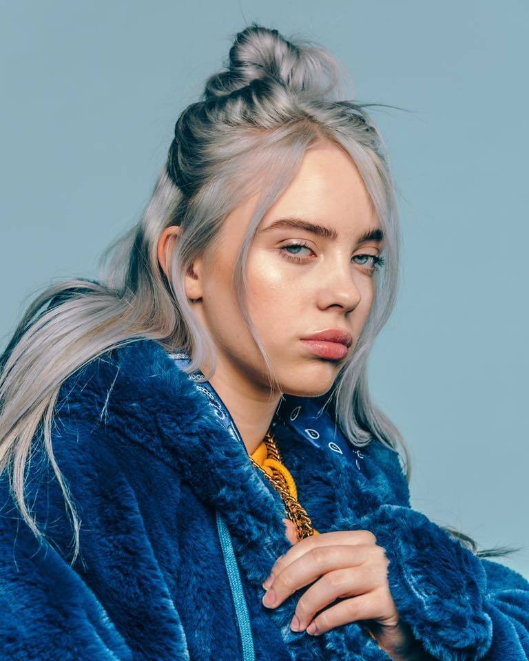 Portrait of a woman with silver hair styled in a top knot, wearing a blue faux fur jacket, against a light blue background.