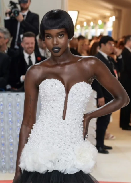 A striking fashion photograph features a Black model in a beaded white gown with dark lipstick, amidst a crowd at a celebrity event.