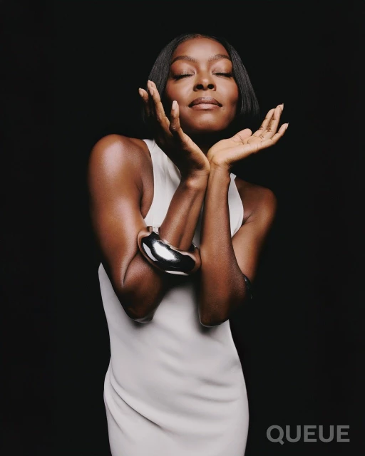 Woman with short black hair and a silver bracelet poses in a sleeveless white dress against a dark backdrop with eyes closed.