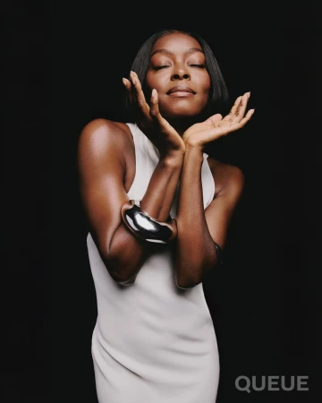 Woman with short black hair and a silver bracelet poses in a sleeveless white dress against a dark backdrop with eyes closed.