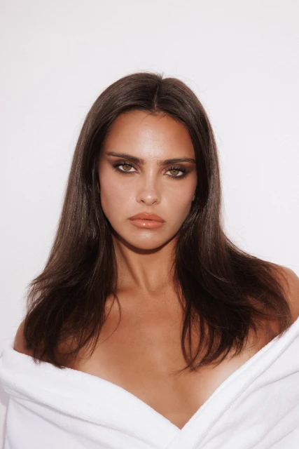 Portrait of a young adult woman with long brown hair and bronze skin wearing a white bathrobe against a white backdrop.