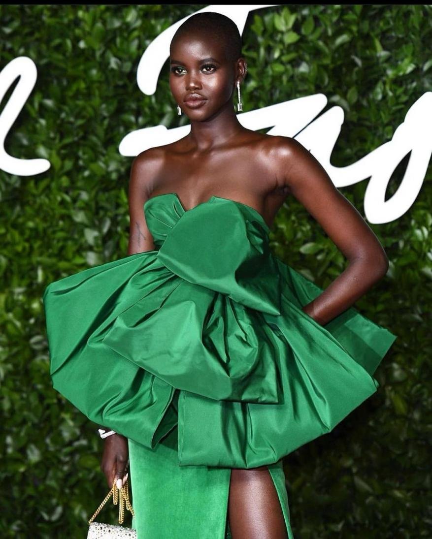 An African American woman with a shaved head poses in a formal floor-length emerald green gown at a fashion event with a leafy background.