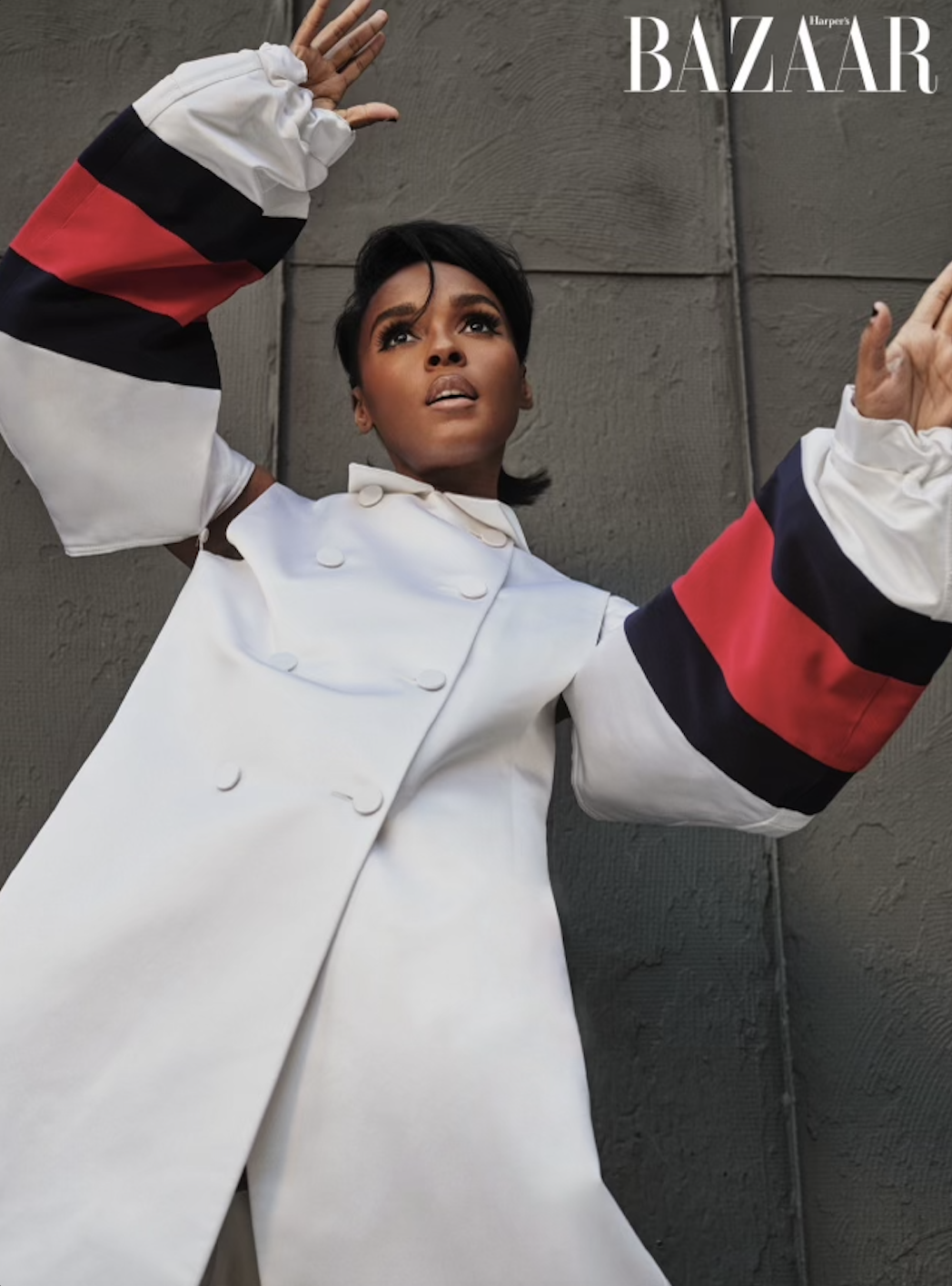 Portrait of adult female in white coat with red and navy sleeves raising arms, looking up against textured grey background with BAZAAR logo.