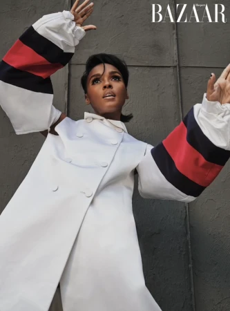Portrait of adult female in white coat with red and navy sleeves raising arms, looking up against textured grey background with BAZAAR logo.