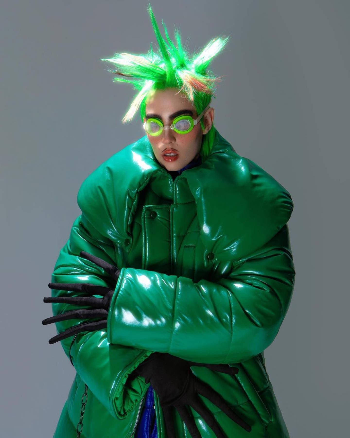 A person with bright green spiky hair, wearing round green glasses and a puffy green coat, poses in a studio with a light gray backdrop.