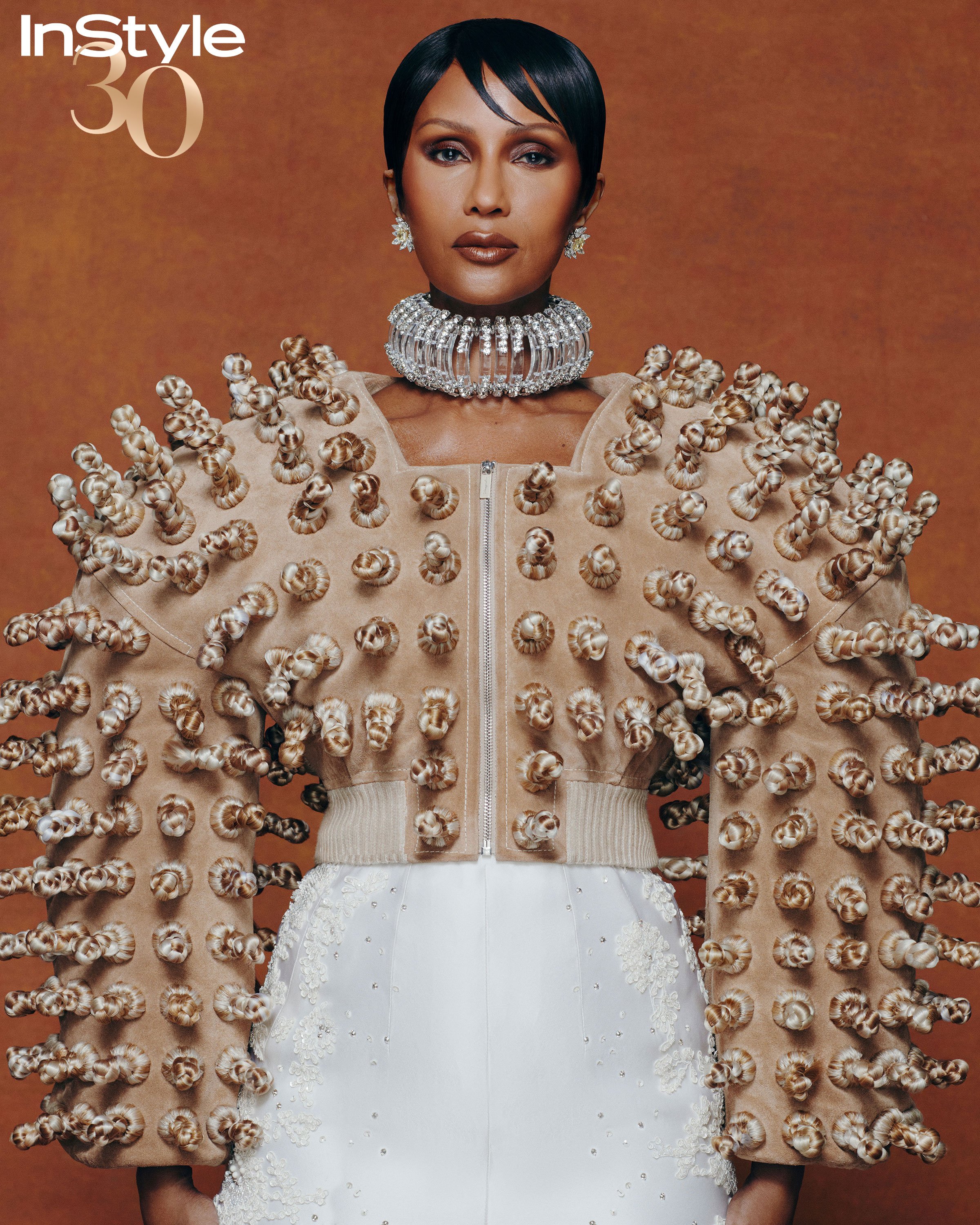 Fashion editorial featuring a Black woman in a sculptural jacket and diamond jewelry, posing against a warm brown background.