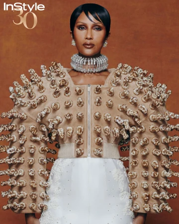 Fashion editorial featuring a Black woman in a sculptural jacket and diamond jewelry, posing against a warm brown background.