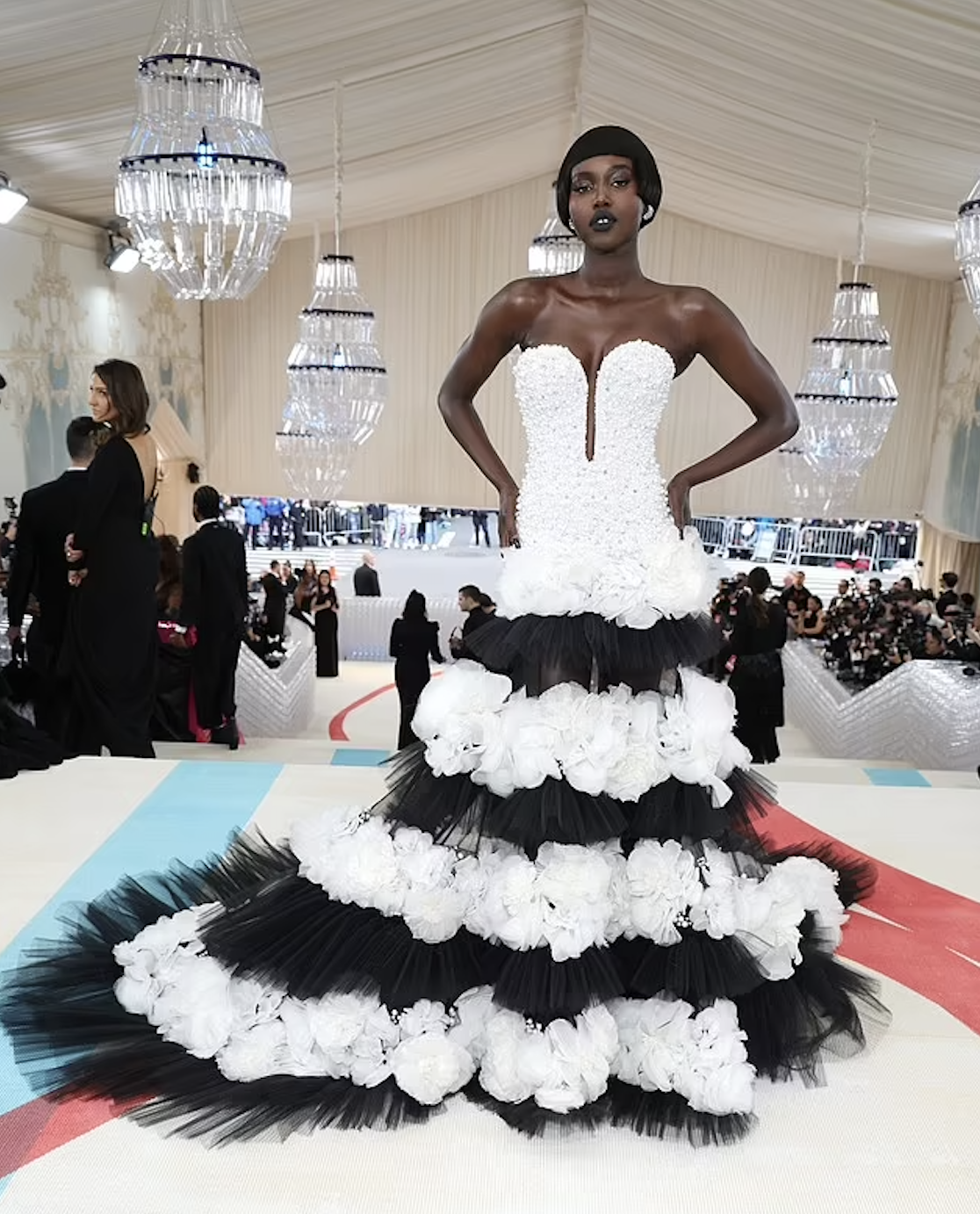 A model showcases a layered black and white gown on a patterned floor at a formal event with photographers and chandeliers.