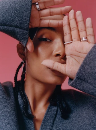 Close-up portrait of a woman framing her face with hands, wearing a grey wool jacket and rings against a pink background.