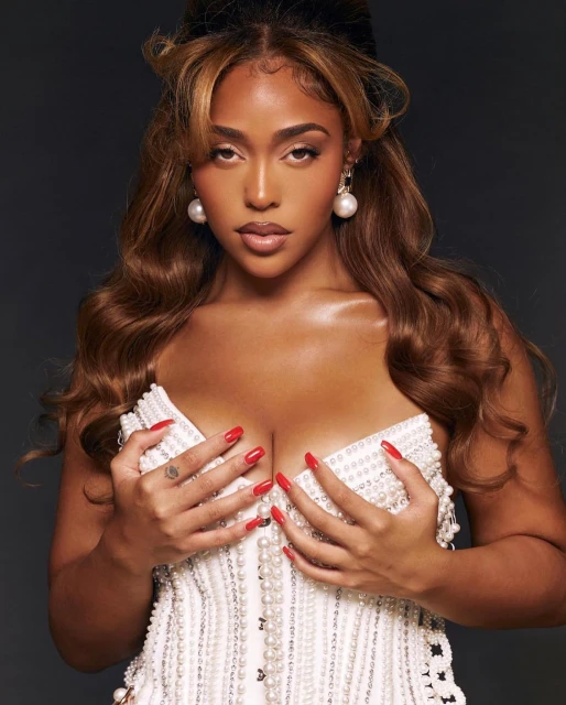 Close-up studio shot of a light-skinned woman with long brown hair wearing a white, pearl-covered top, and large pearl earrings.