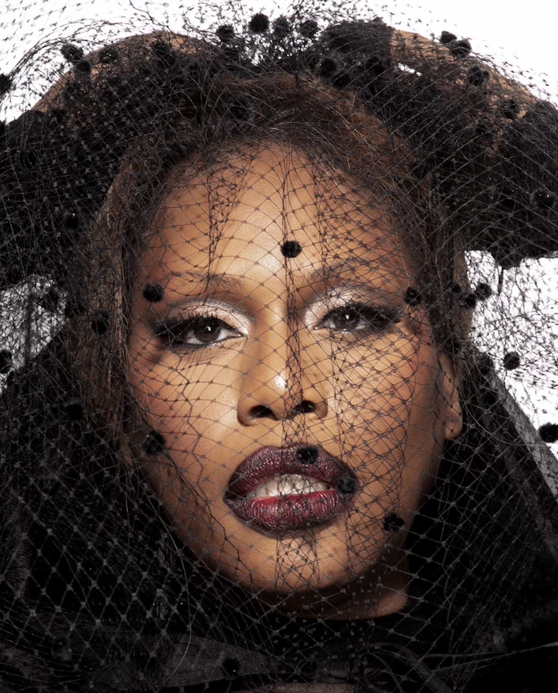 Close-up portrait of an adult female model with brown hair and dark lipstick posing in a black, dotted mesh veil on a white background.