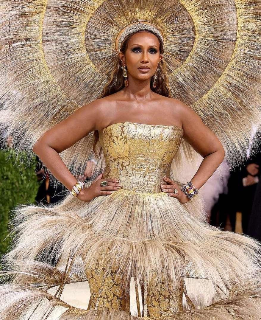 Iman poses in a gold strapless gown and headpiece resembling feathers on the red carpet at an event, hands on her waist.