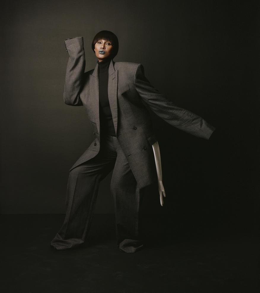 Fashionable adult female poses in a gray oversized suit in a studio setting with a dark backdrop, creating a dramatic fashion portrait.