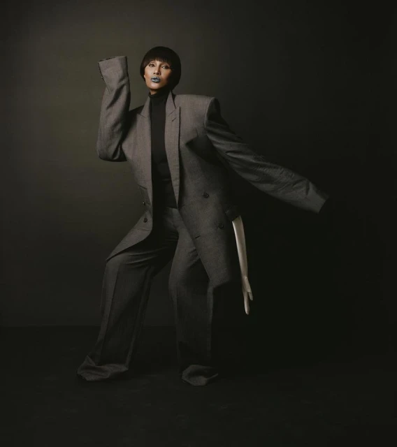 Fashionable adult female poses in a gray oversized suit in a studio setting with a dark backdrop, creating a dramatic fashion portrait.