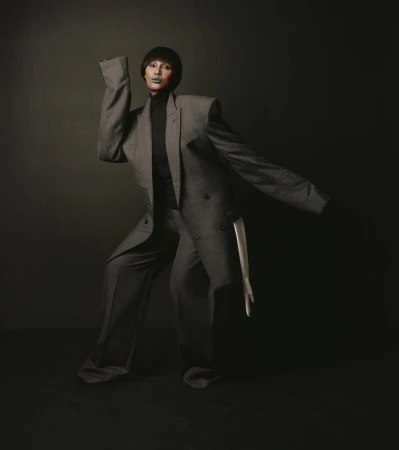 Fashionable adult female poses in a gray oversized suit in a studio setting with a dark backdrop, creating a dramatic fashion portrait.
