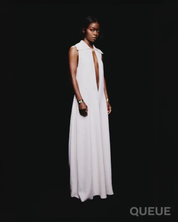 Black woman wearing a long white sleeveless dress and gold cuff bracelets against a black background.