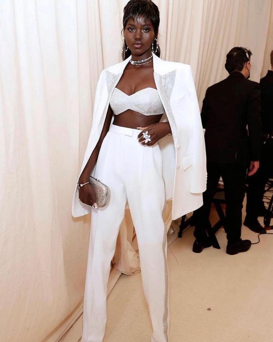 Elegant woman in a white pantsuit, bejeweled bra top, and silver clutch, standing in an indoor setting with soft, ambient lighting.