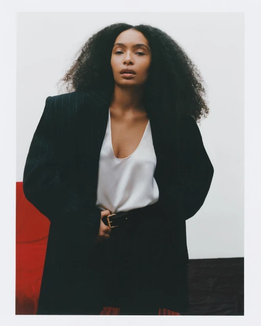 Portrait of a woman with an afro wearing a black blazer and a white shirt in front of a white, red, and black backdrop.