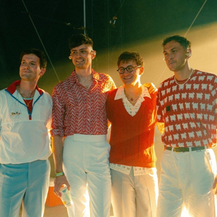 Group portrait of four young adult men in vibrant patterned shirts and white pants, standing together indoors in warm lighting.