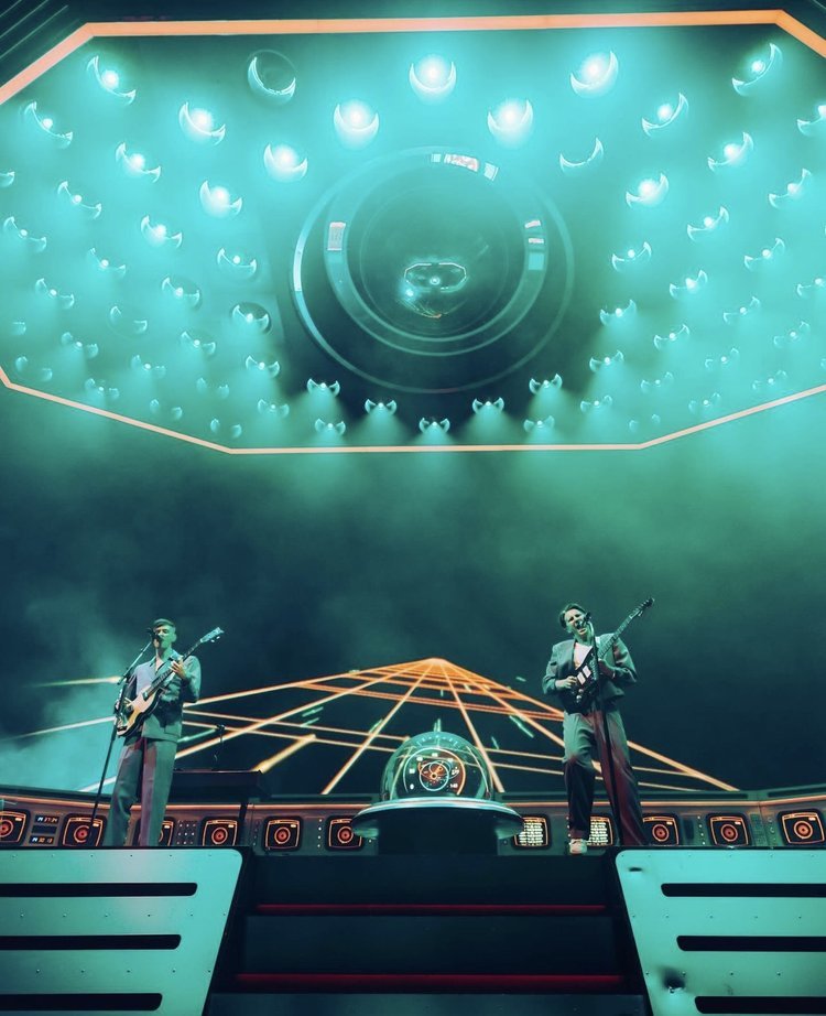 Two musicians perform on an illuminated stage with instruments and lighting design, wearing suits and playing guitars under teal lighting.