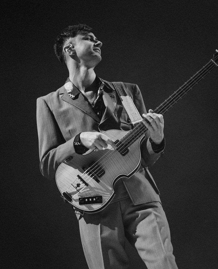 Monochromatic close-up of a musician in a suit playing an electric bass guitar, set against a dark background, bathed in stage lighting.