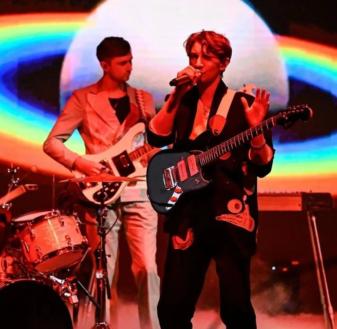 Singer performs on stage with guitarist in background, both in suits. Stage lights and screen illuminate the music performance.
