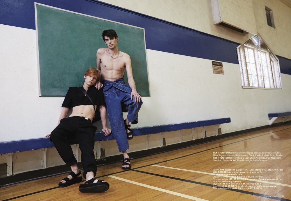 Two shirtless men pose in a gymnasium, one seated on a blue ledge in a short black top and the other standing in denim pants.