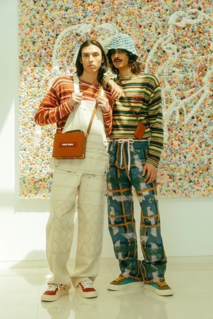 Two men pose indoors against colorful art. One wears a striped shirt, blue hat, and patterned pants. The other has a box bag.