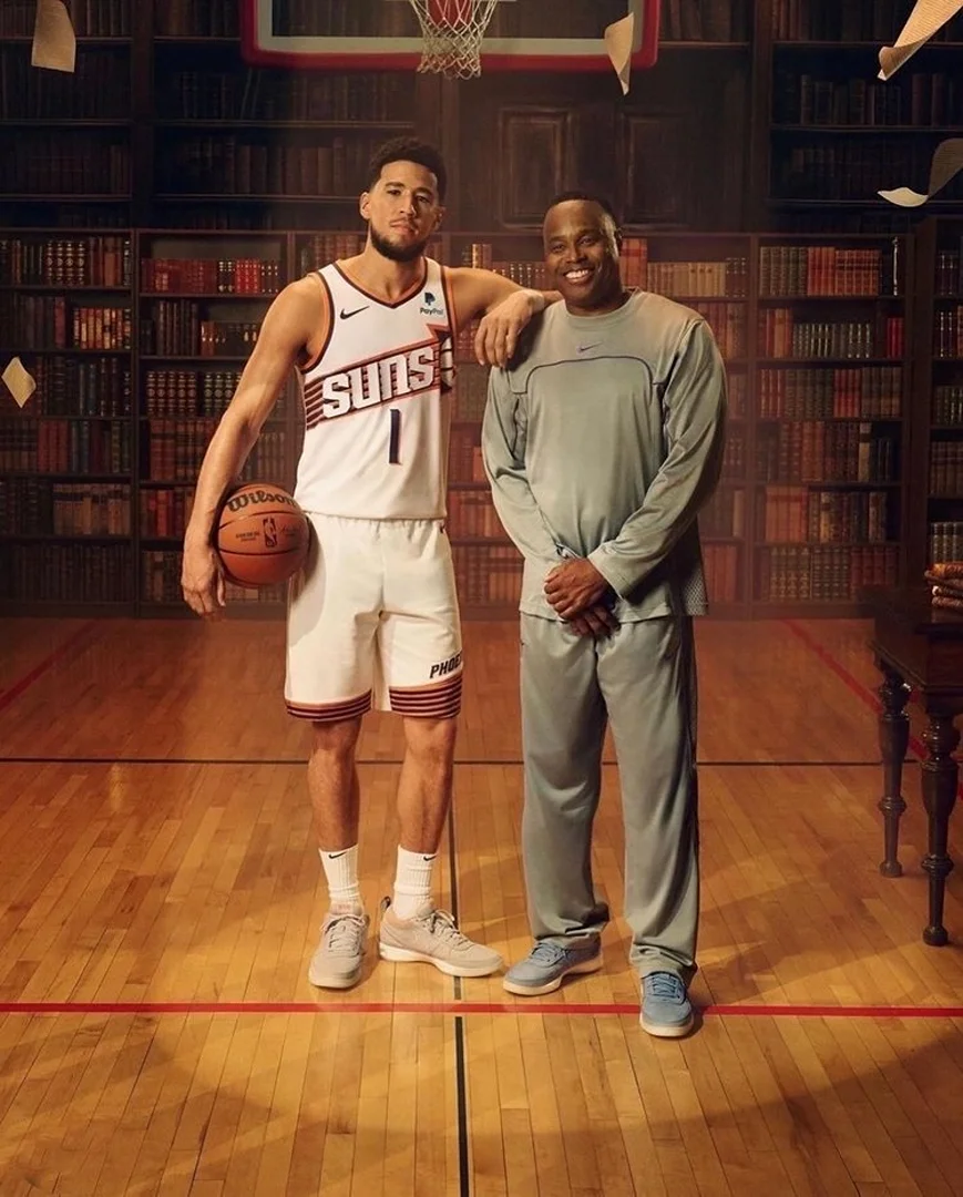 Two men stand on a basketball court in front of a bookshelf: one wearing a Suns jersey holding a basketball, the other in grey athletic wear.