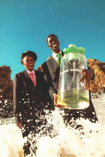 Two smiling people in suits stand in ocean surf holding an oversized perfume bottle against a bright blue sky and rocky shoreline.