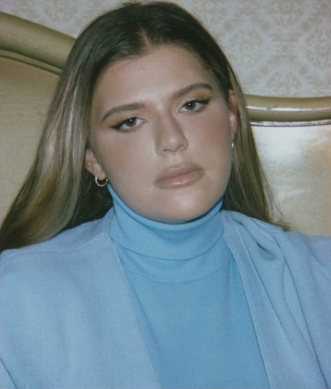 Close-up portrait of an adult woman with long hair wearing a light blue turtleneck and jacket, posing against a patterned backdrop.