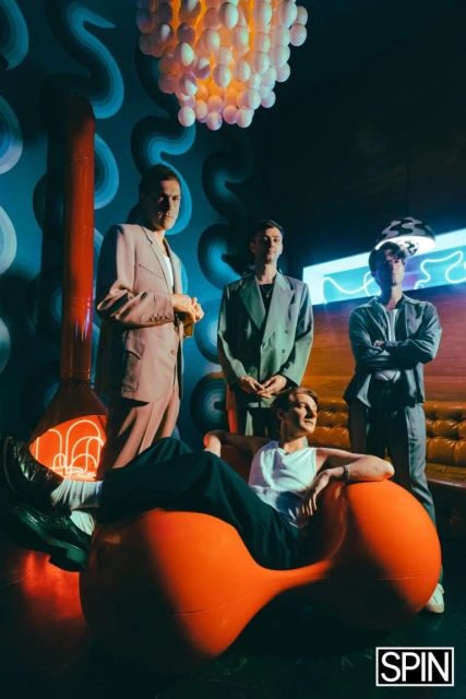 Group shot of four adult men in a stylish interior with an orange chair, mood lighting, and fashionable suits, reminiscent of a band portrait.