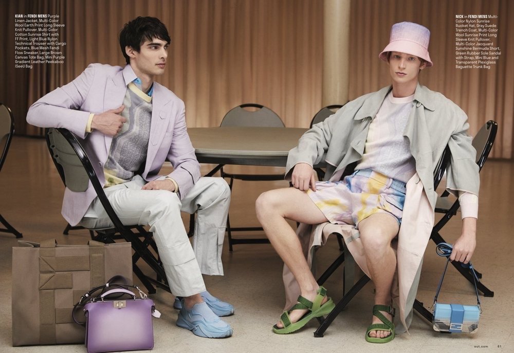 Two adult male fashion models pose sitting in chairs with designer apparel and bags in an indoor setting with tables and neutral walls.