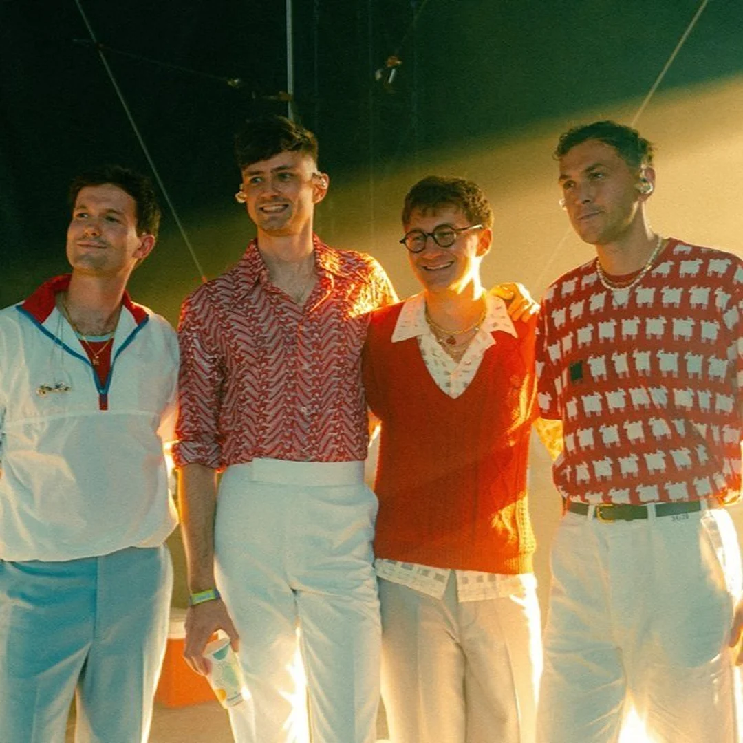 Group of four young adult men pose in patterned shirts and pants in an indoor setting with a stage-like background.