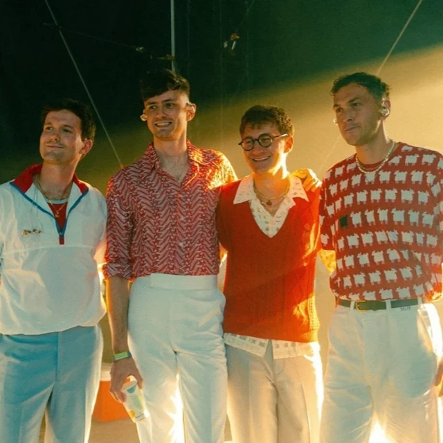 Group of four young adult men pose in patterned shirts and pants in an indoor setting with a stage-like background.