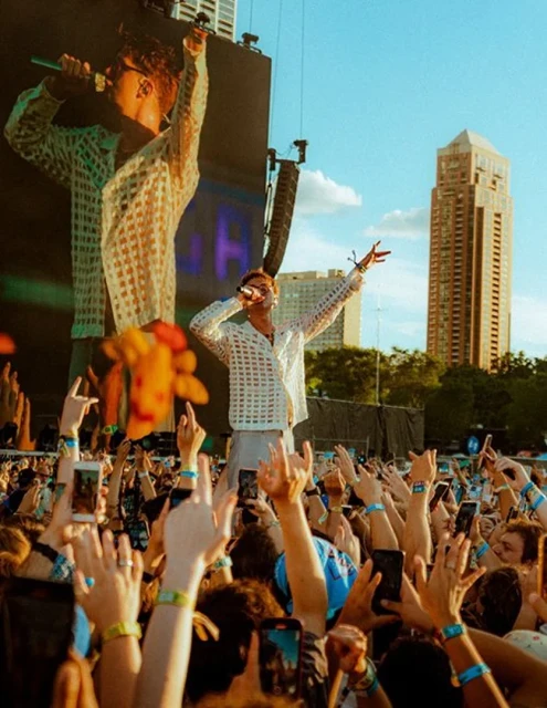 Male singer performing on stage during an outdoor concert with a large crowd of people holding up their cell phones to take pictures.