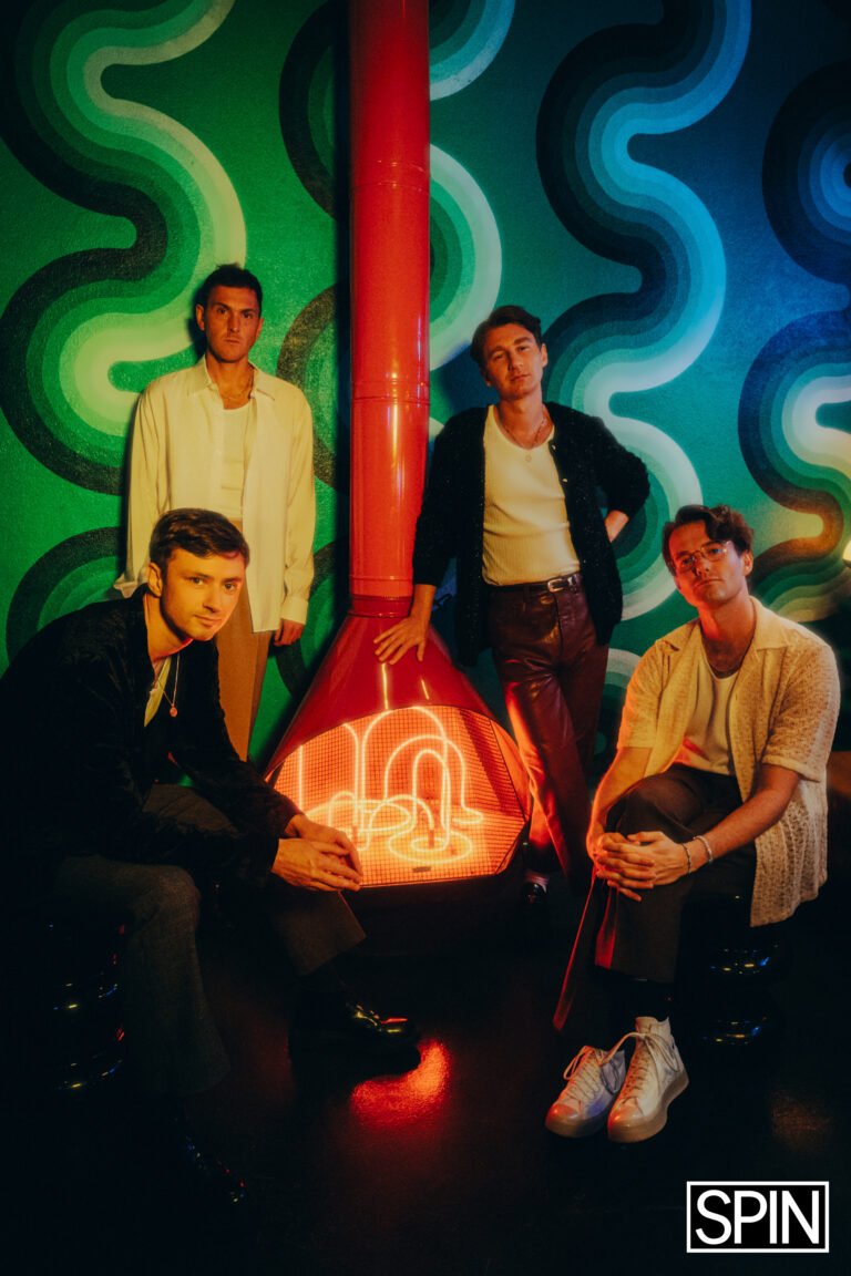 Group portrait of four young adult men in casual outfits standing near a retro-style orange fireplace against a green patterned wall.