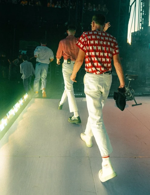 Four male performers in matching outfits walk on stage during a live music concert, captured from a medium shot behind the performers.