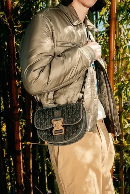 Person wearing a green jacket and khaki pants carrying a Dior saddle bag, standing in front of a bamboo forest background.